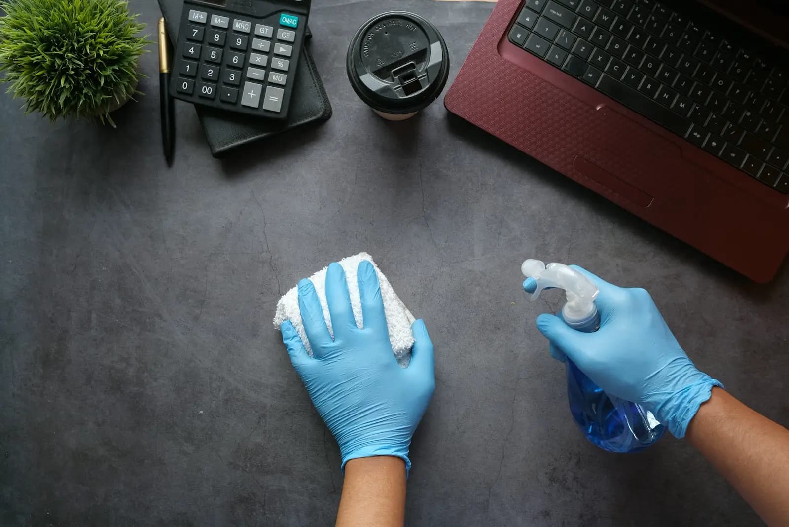 Mãos com luvas a limpar uma secretária de escritório com pano e pulverizador.